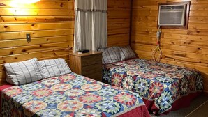A cabin-style room with two beds, a nightstand, a window with curtains, and an air conditioning unit.