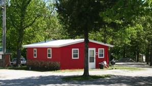 A red single-story building with a white door and windows, surrounded by trees and a gravel driveway. - Cabin 6 - Reel and Trigger (Tom Township)