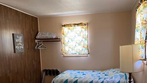 A room with two beds, wooden paneling on one wall, a window with a floral curtain, and a ceiling light. - Cabin 6 - Reel and Trigger (Tom Township)