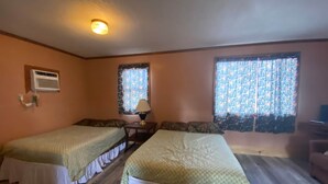 A room with two beds, a small table with a lamp, a window with patterned curtains, and a ceiling light. - Cabin 8 - Reel and Trigger (Tom Township)