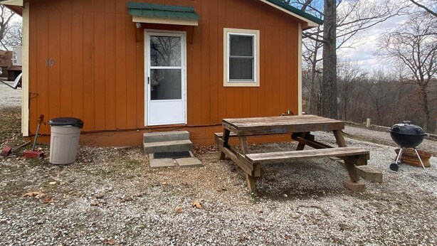 Une petite cabane en bois avec un toit vert, une porte blanche et une fenêtre. On y trouve à l’extérieur une table de pique-nique et un barbecue.