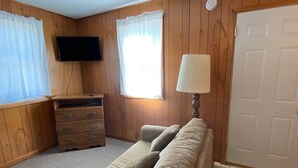 Une pièce aux murs recouverts de panneaux de bois, un canapé beige avec des coussins, une commode en bois avec des tiroirs, une lampe, un téléviseur fixé au mur et une porte.