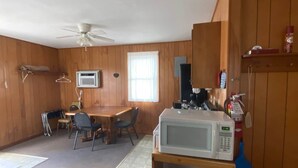 Une cuisine compacte aux murs en bois, un coin repas avec une table et des chaises, un four à micro-ondes posé sur une étagère et un ventilateur de plafond.