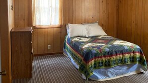A bedroom with wooden paneling, a bed with a colorful quilt, a window with curtains, and a small wooden cabinet. - Eagle's Nest - Reel and Trigger (Tom Township)
