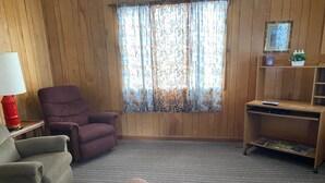 A cozy room with a wooden paneled wall, a purple armchair, a brown sofa, a small table with a lamp, and a TV stand with a remote. - Eagle's Nest - Reel and Trigger (Tom Township)
