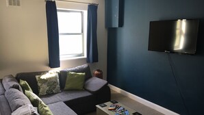 A living room with a sectional sofa, a coffee table with magazines, a flat-screen TV mounted on the wall, and a window with curtains. - Hurricane - 2 bed apartment (Friskney)