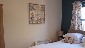 A bedroom with a bed, a radiator, a nightstand with a lamp, and a wall-mounted artwork. - Spitfire - 2 bedroom apartment (Friskney)