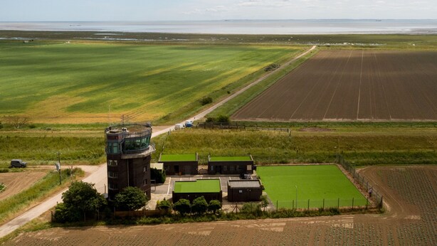An aerial view of a farm with green fields, a building with a green roof, and a body of water in the distance. - Spitfire - 2 bedroom apartment (Friskney)