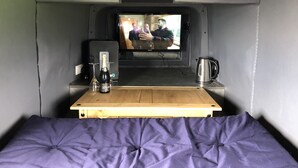 A compact living space with a television mounted on a wall, a small table with a bottle and glasses, and a kettle on the side. - Glamping pod helicopter (Friskney)