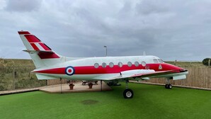 Uma pequena aeronave estacionada numa área gramada, com uma cerca de madeira ao fundo.