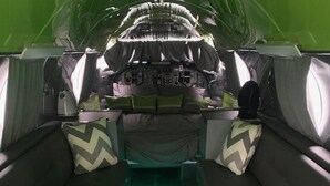 Interior de aeronave com corredor central, iluminação verde e painel de controle na frente.