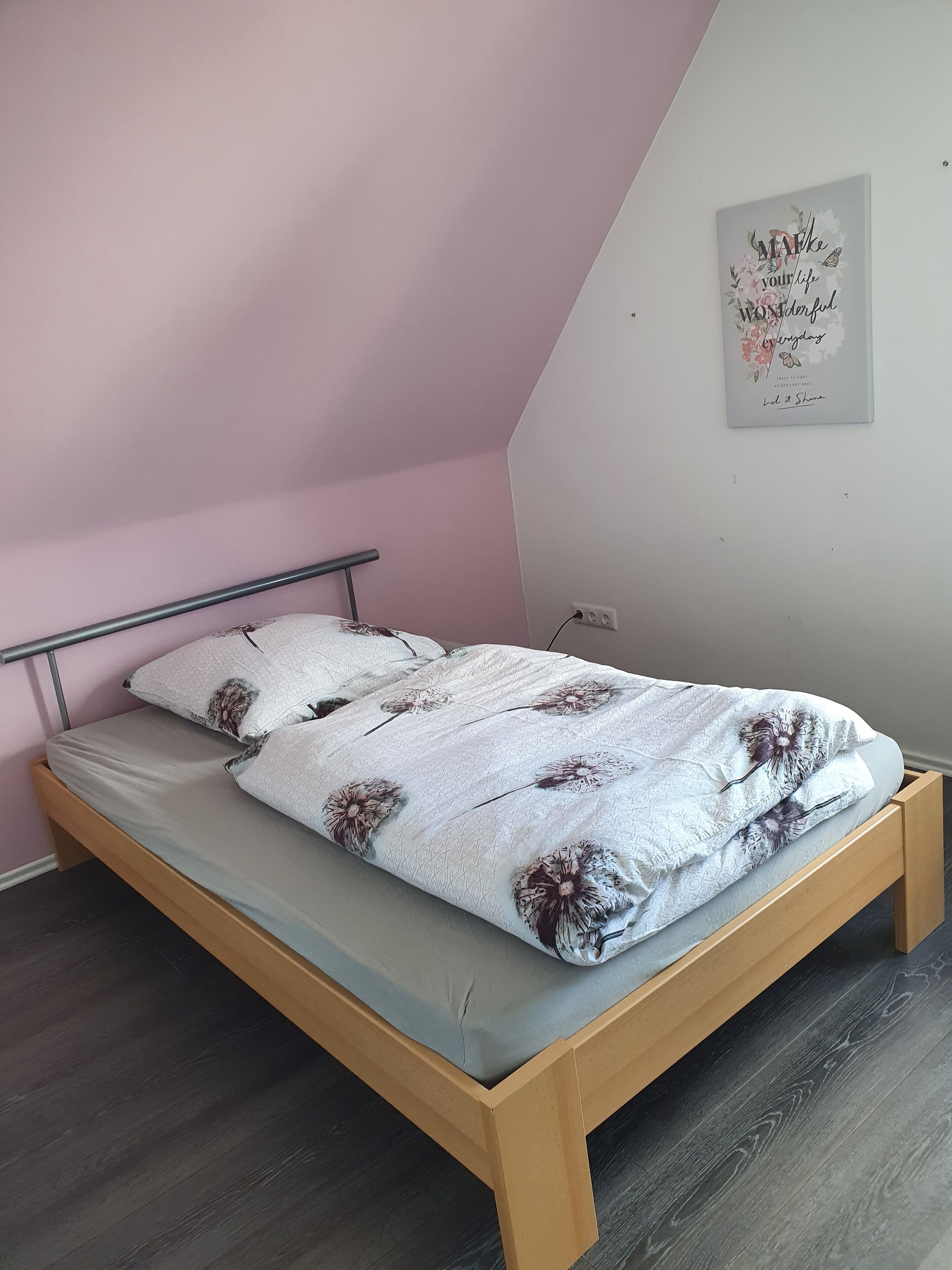Ein Einzelbett mit Holzrahmen, grauer Matratze, einem Metall-Kopfstück und einer Bettdecke mit Blumenmuster.