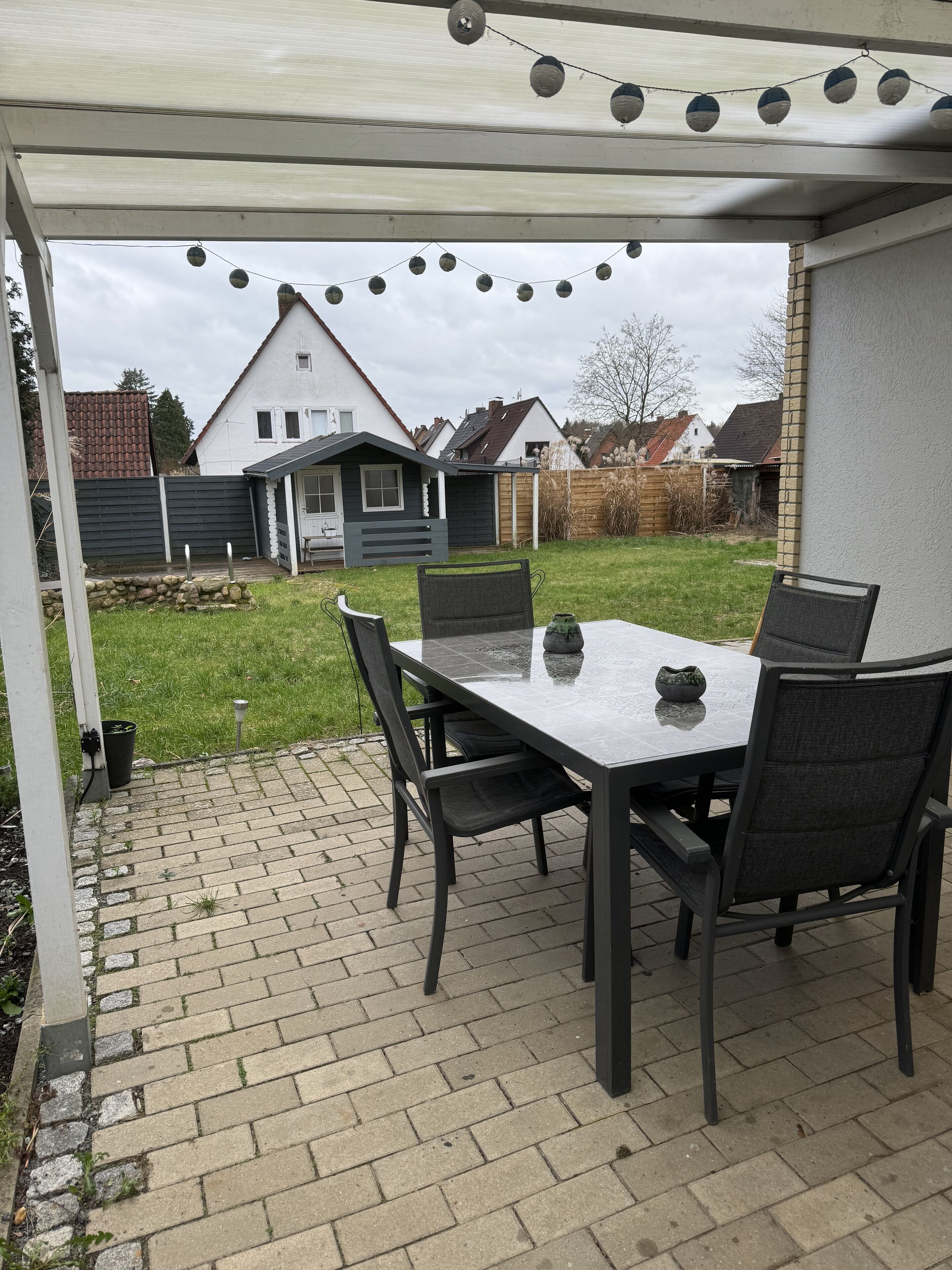 En uteplats med ett bord och stolar, en ljusslinga och utsikt över ett hus i bakgrunden.