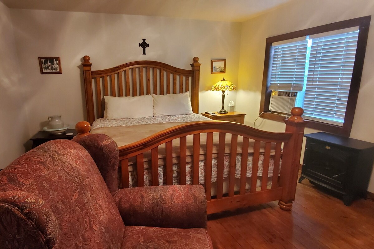 Une chambre à coucher avec un lit en bois, une table de chevet, une lampe, une chaise, une commode et une fenêtre avec des stores.
