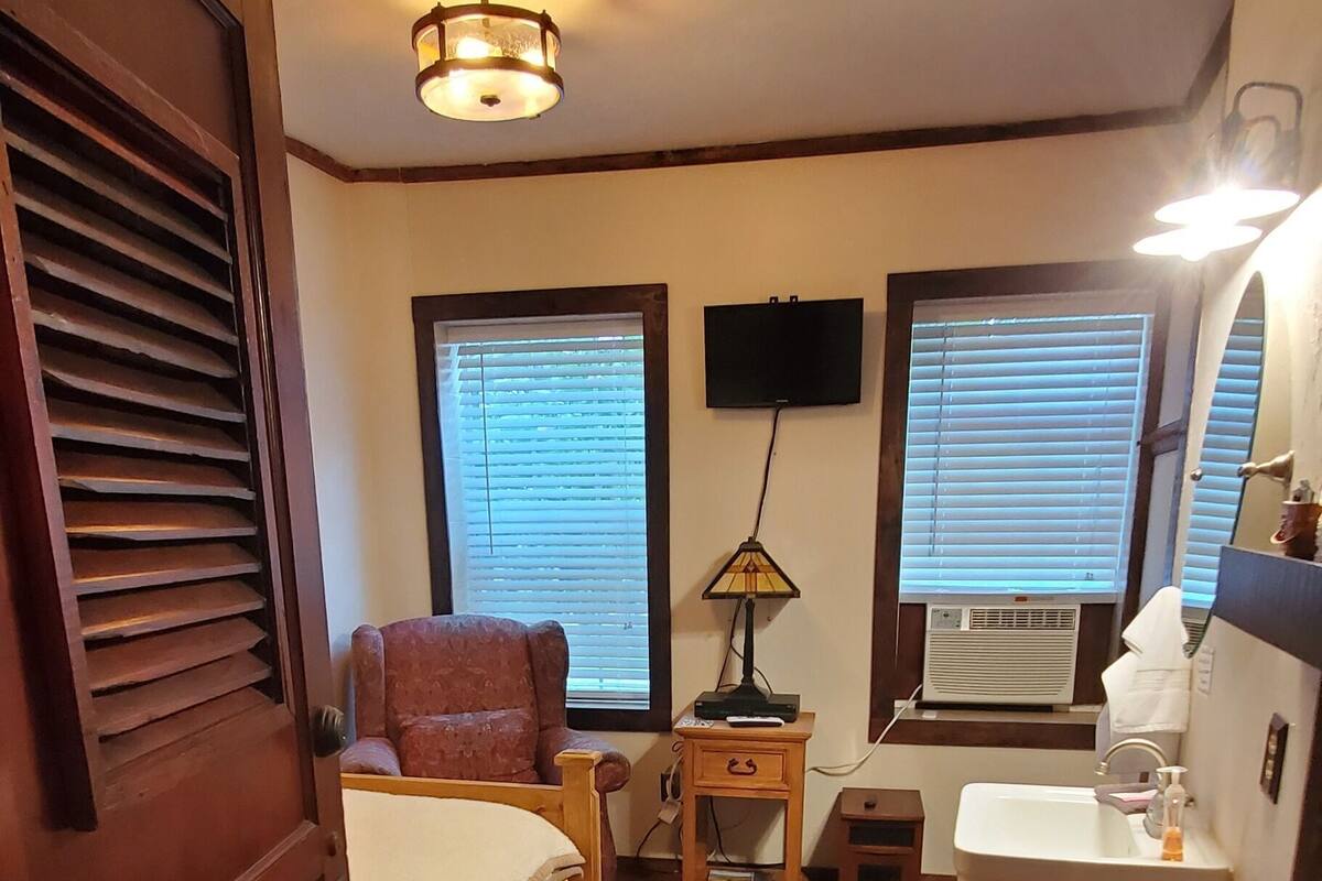 Una habitación con cama, una silla, una mesita de noche con lámpara, un lavabo y un televisor montado en la pared.