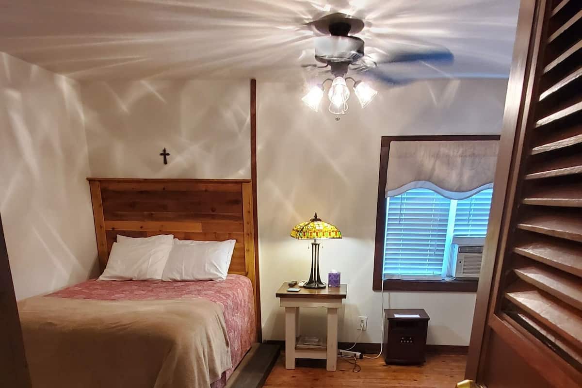 Un dormitorio con una cama de madera, una mesita de noche con lámpara, un ventilador de techo y una ventana con persianas.