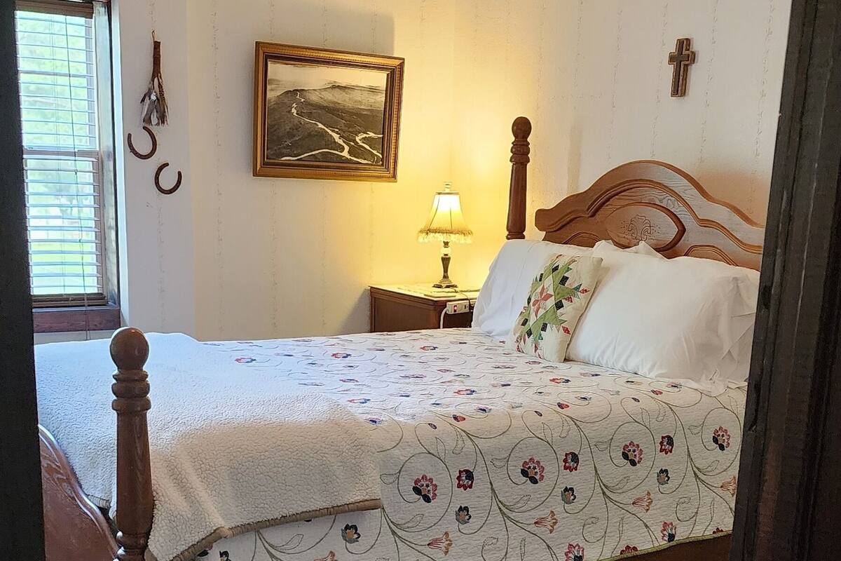 Un dormitorio con cama, ventilador de techo, mesita de noche, lámpara, un cuadro enmarco, una cruz en la pared y ventana con persianas.