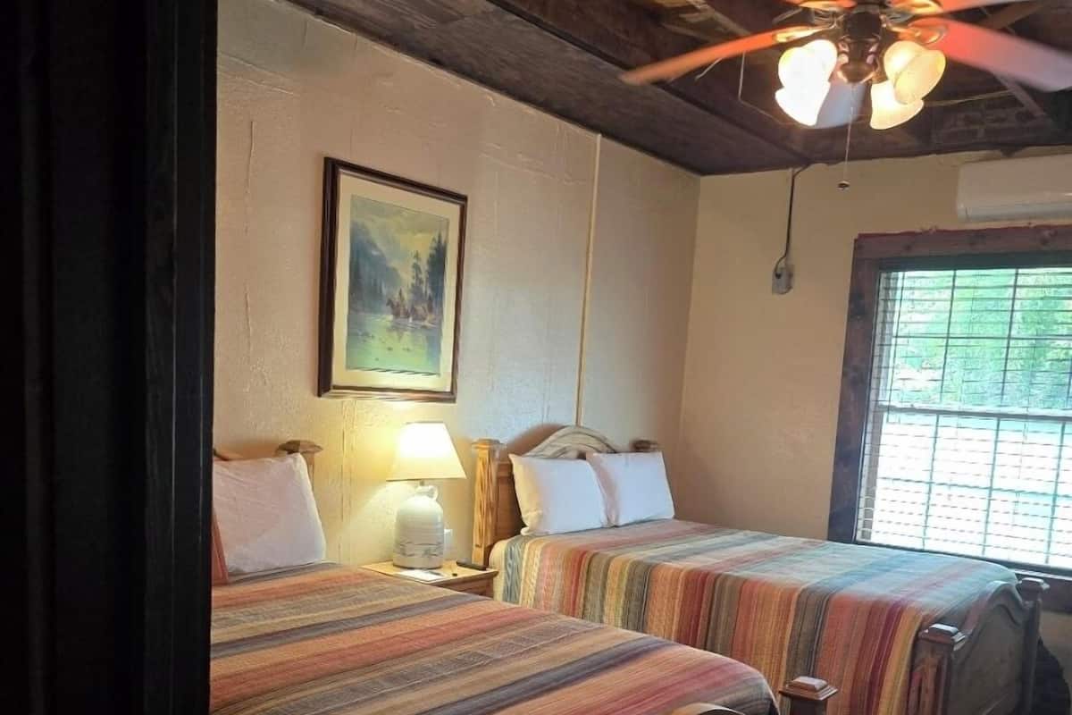 Una habitación con dos camas de madera, un ventilador de techo, un cuadro en la pared y una ventana con persianas.