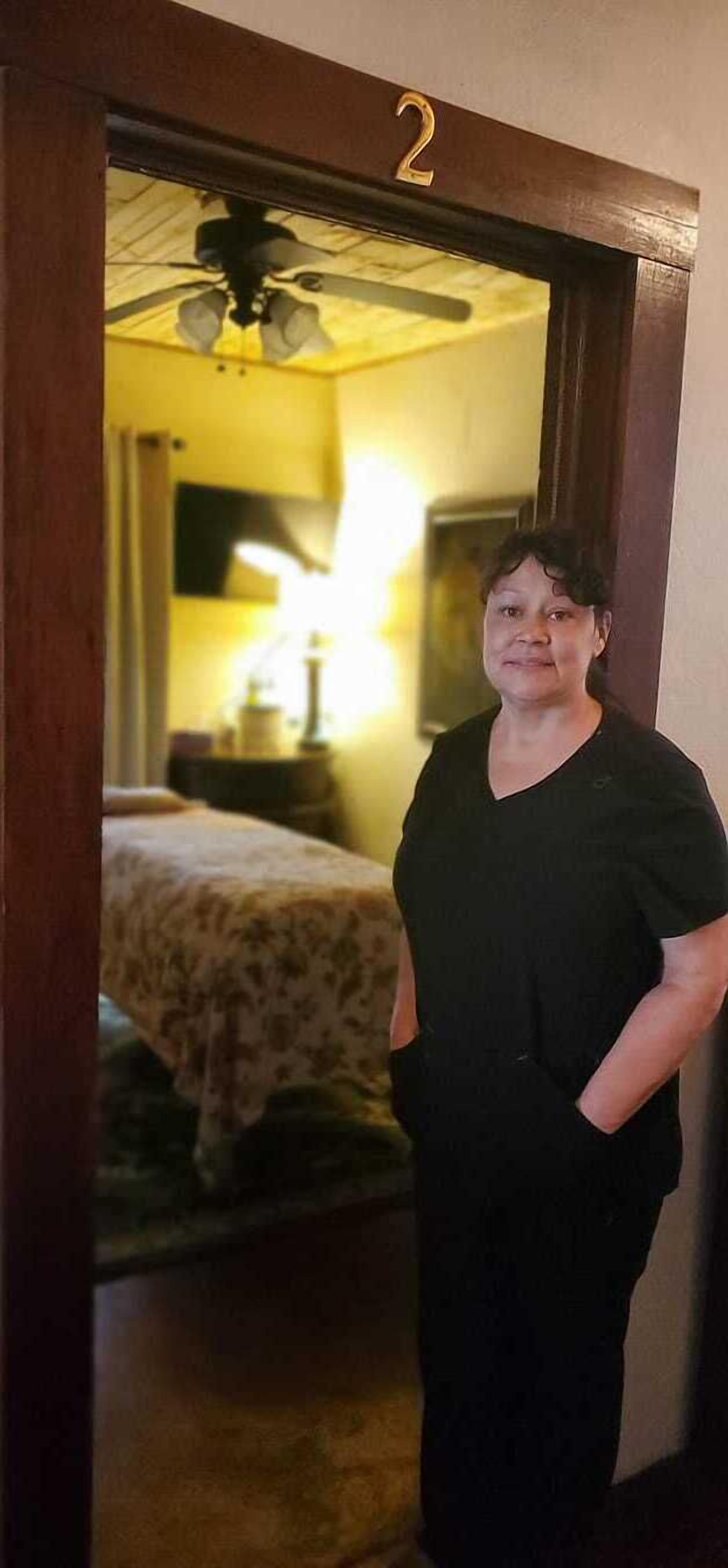 A person standing in a doorway with a room visible in the background. The room has a bed with a floral bedspread, a lamp, and a ceiling fan.
