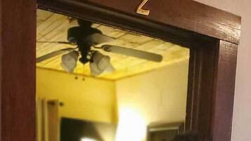 A person standing in a doorway with a room visible in the background. The room has a bed with a floral bedspread, a lamp, and a ceiling fan.