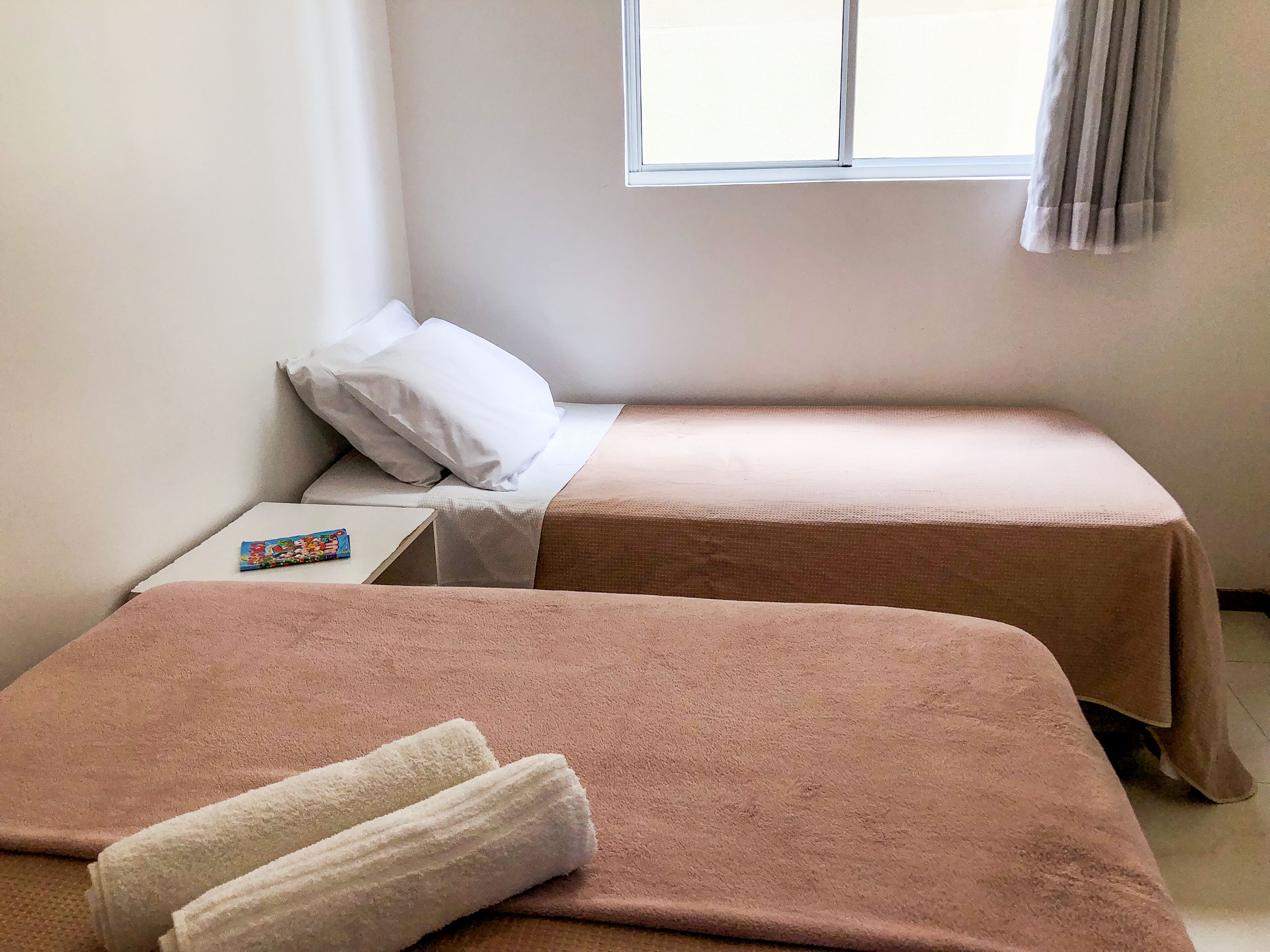 A room with two beds, each with a pink bedspread and white pillows. There is a small table with a book and a remote control.