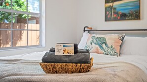 A neatly made bed with a basket containing a towel and a card, a painting on the wall, and a window with a view of greenery and a house.