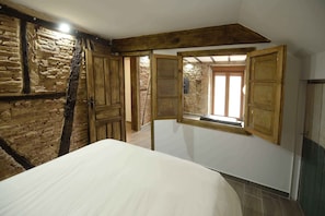 A bedroom with a bed, wooden beams, stone wall, and a window with wooden shutters.