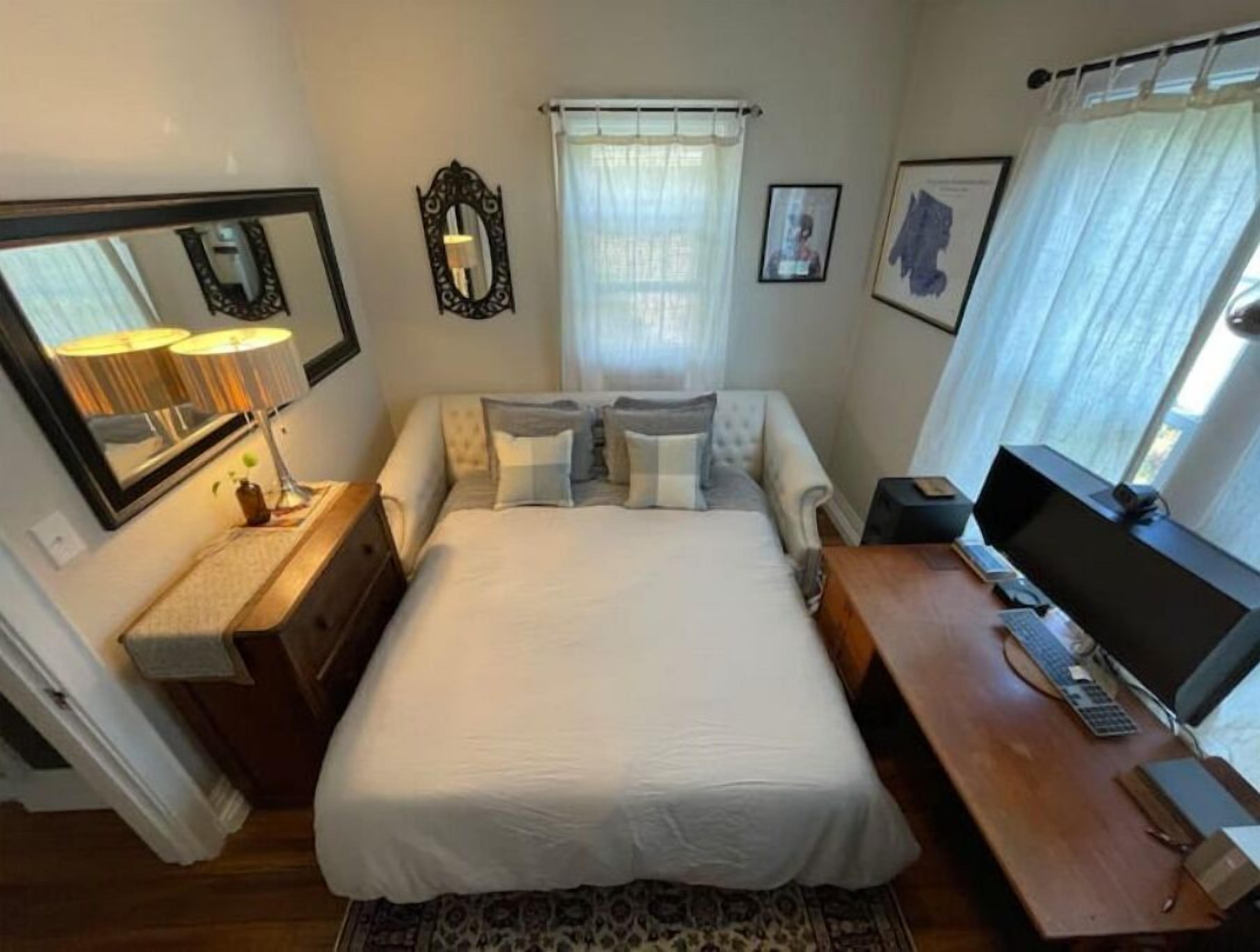 A neatly arranged bedroom with a bed, bedside table, lamp, mirror, and a desk with a computer.