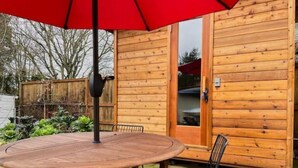 A wooden cabin with a round wooden table and metal chairs, a red umbrella, and a wooden door. - 2Bd Bungalow with backyard sauna, by Alberta Arts (Portland)