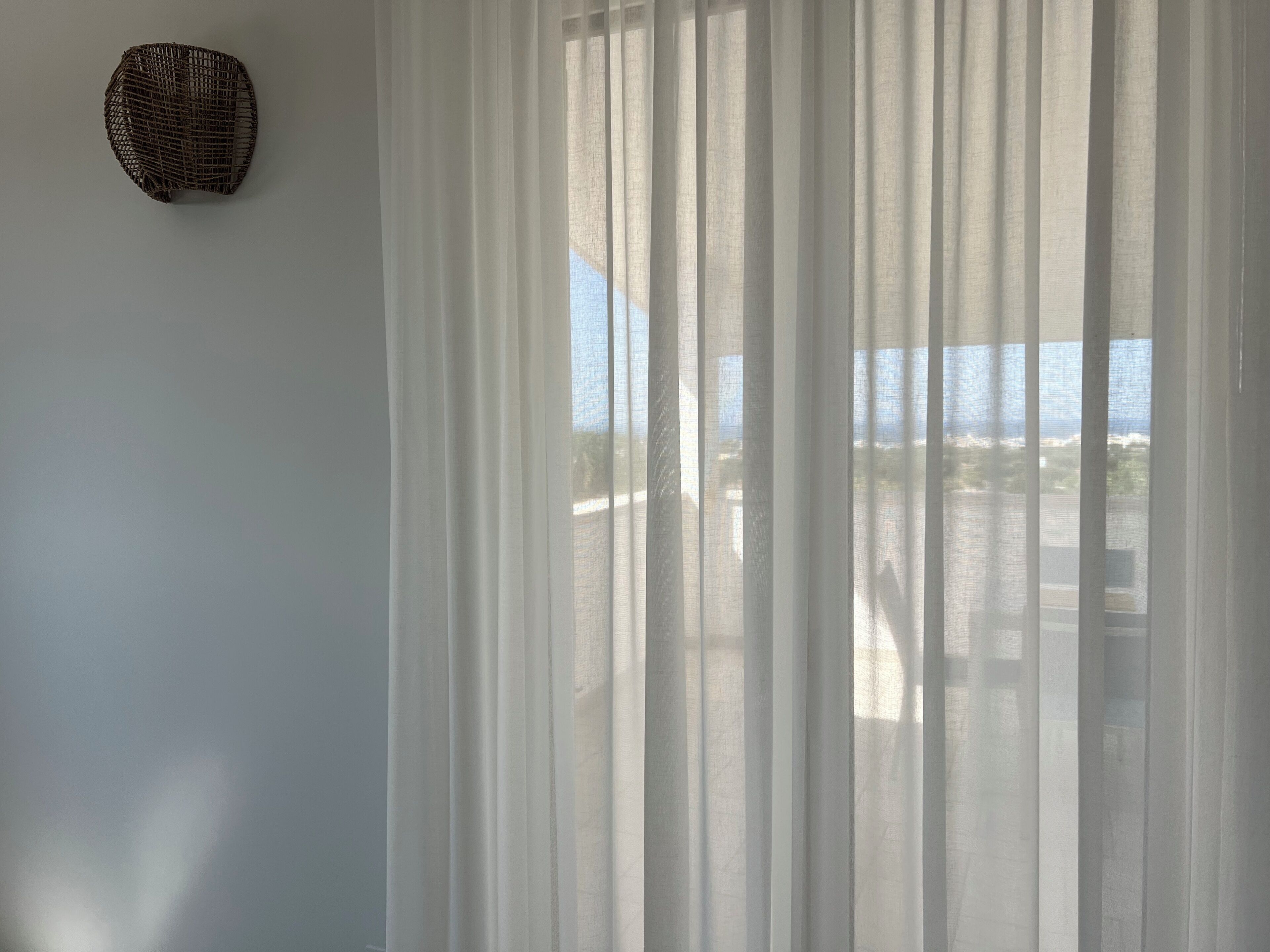 A room with sheer white curtains, a wicker wall decoration, and a view of the sea.