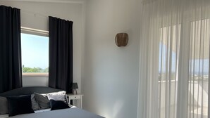 Une chambre à coucher avec un lit, une table de chevet, une lampe et une vue sur l’extérieur à travers des rideaux légers.