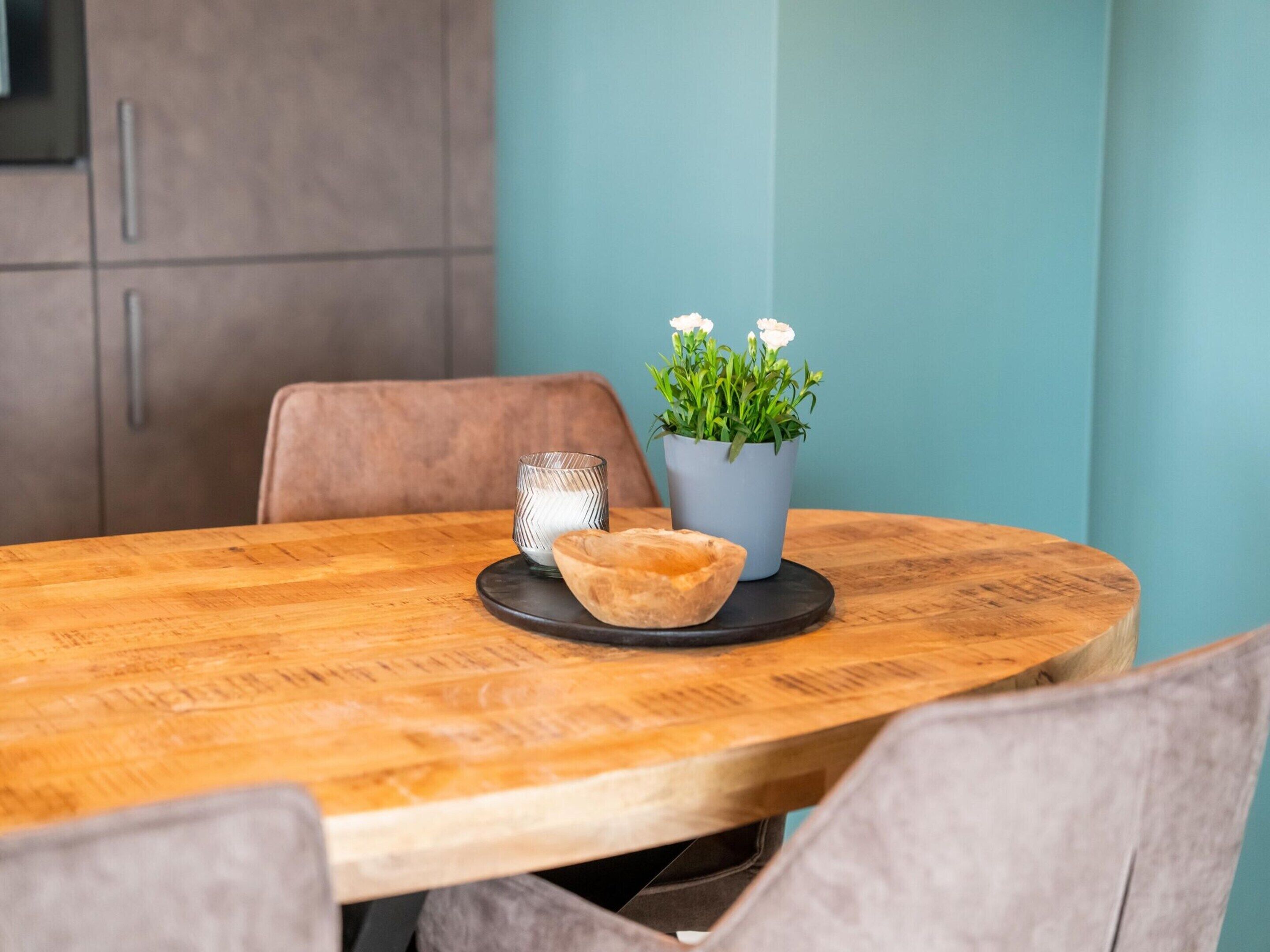A wooden dining table with a bowl and a glass on a black coaster, a potted plant with white flowers, and a brown chair.