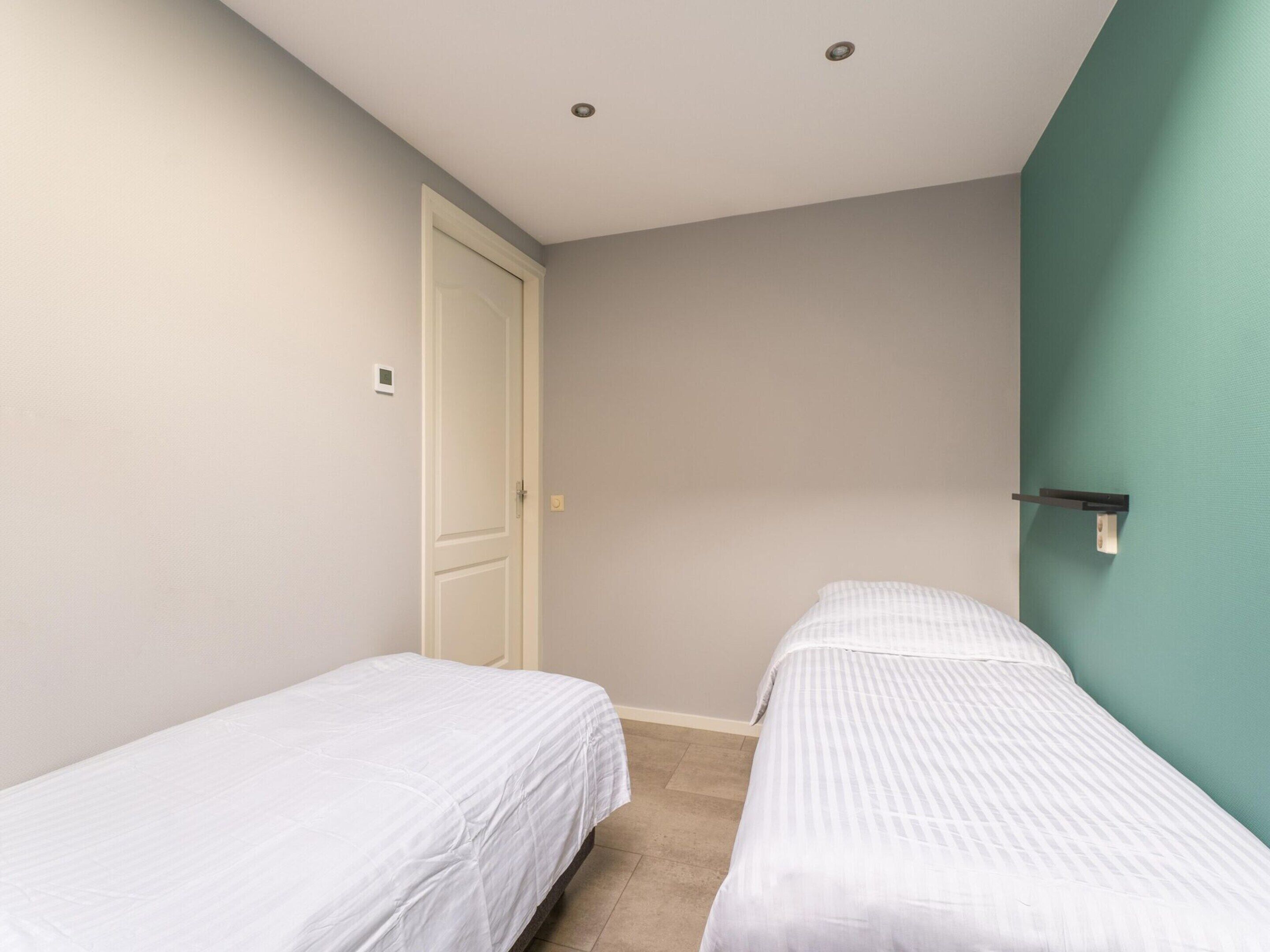 A small, clean room with two single beds, a door, and a light switch on the wall.