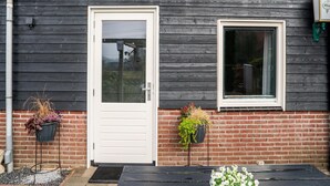 A building with a dark wooden exterior, a white door, and a window. There are two planters with plants on the ground. - Charming House With Garden (Hellendoorn)