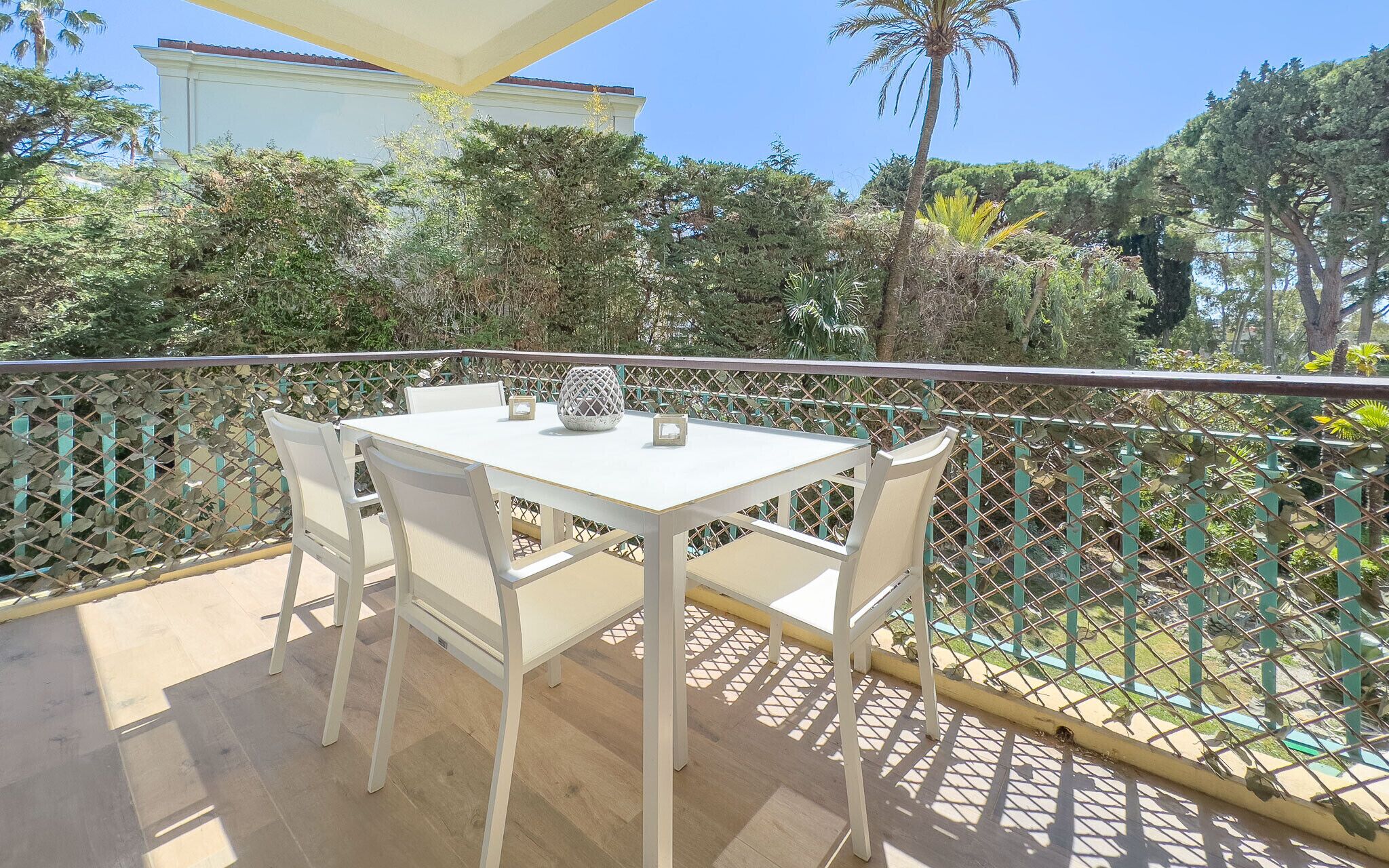 Ein Balkon mit weißem Tisch und Stühlen, einer Dekovase und Blick auf umliegendes Grün sowie ein Gebäude.