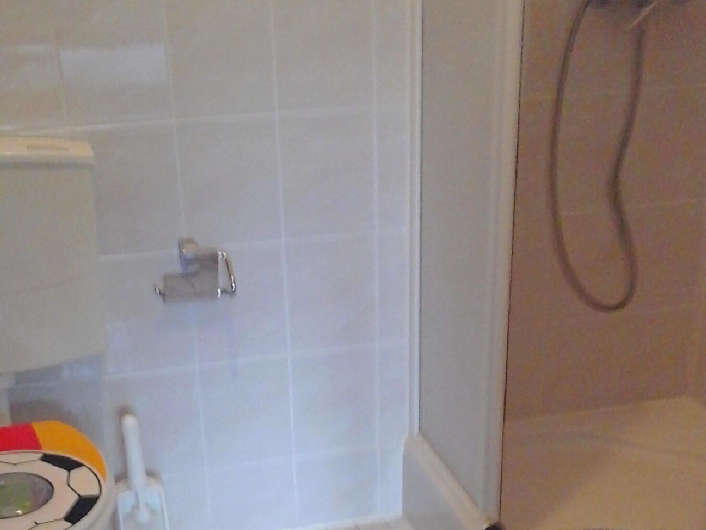 Une salle de bain avec des carreaux blancs, des toilettes, une douche et un objet coloré sur le sol.