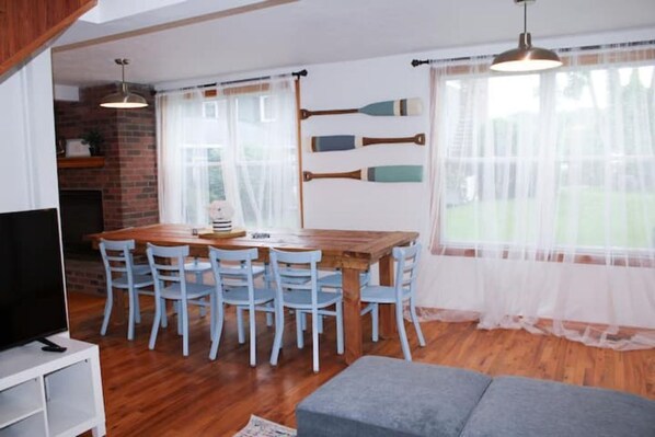 Un comedor con una mesa de madera y seis sillas, un televisor sobre un soporte blanco y un sofá gris.