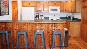 Una cocina con armarios de madera, un microondas blanco y un lavamanos de acero inoxidable. Hay tres taburetes y un cubo de basura.