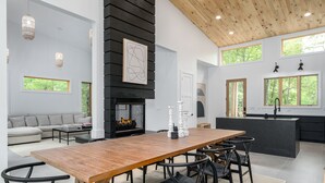 Uma sala de estar moderna com uma mesa de jantar de madeira, cadeiras pretas, lareira e uma cozinha com uma janela grande.