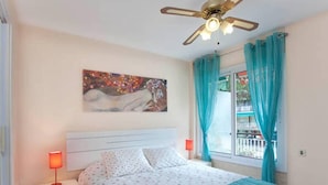 Une chambre à coucher comprenant un lit, une table de chevet, un ventilateur de plafond, une fenêtre avec des rideaux et un tableau accroché au mur.