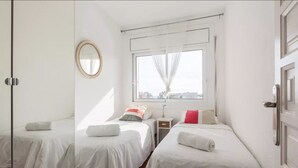 Habitación de hotel con dos camas, una ventana con cortinas translúcidas, una mesita de noche, un espejo y vistas a la ciudad.