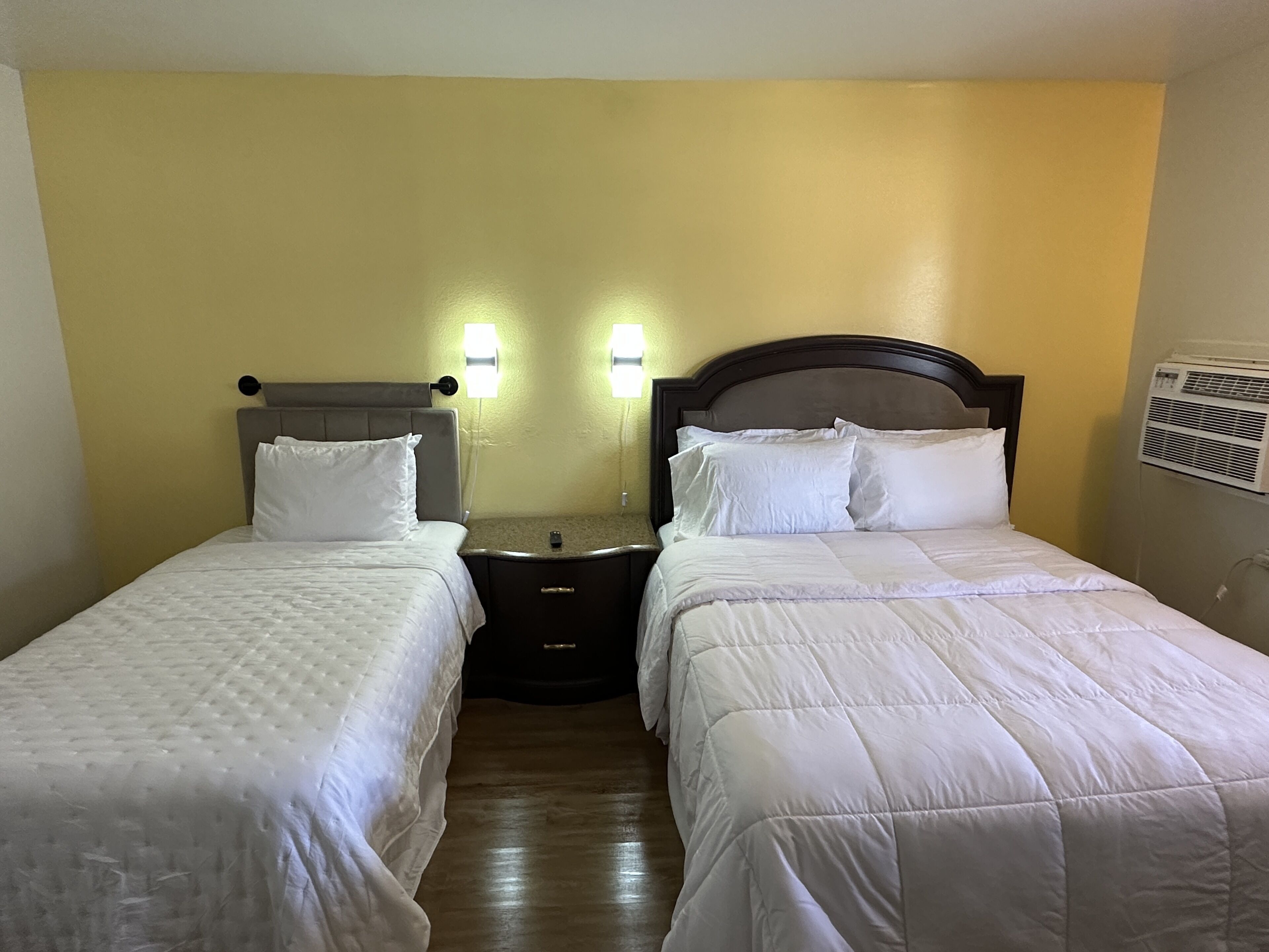 A hotel room with two single beds, a nightstand, and a wall-mounted air conditioner.