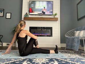 A person sitting on a patterned rug in a room with a fireplace, a television, and a chair with a blue cushion.