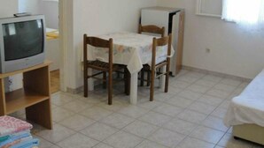 Een kleine kamer met een televisie op een standaard, een tafel met stoelen, een bed en een raam met gordijnen.
