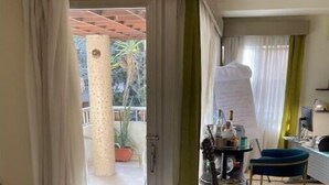 Een kamer met een piano, een gitaar en een bureau met een computer. Een deur leidt naar een buitenruimte met een houten pergola en potplanten.