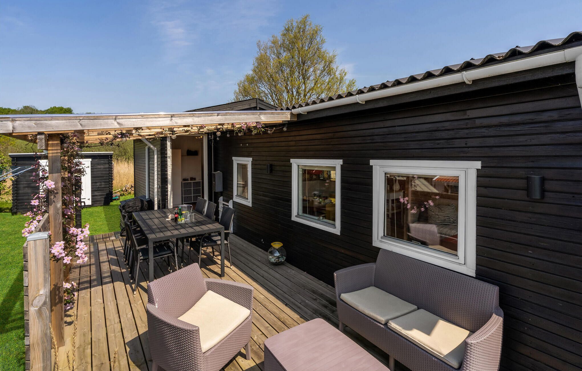 Puinen terassi ulkokalusteineen, kukilla koristeltu pergola ja tumma puinen rakennus, jossa on ikkunat.
