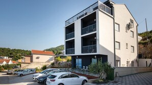 Un edificio de varios pisos con balcones, un estacionamiento con varios coches y un paisaje montañoso al fondo.