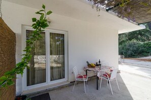 A covered outdoor seating area with a table and chairs, a window with curtains, and a view of greenery. - Apartments Skurla - One Bedroom Apartment With Terrace (Mljet)