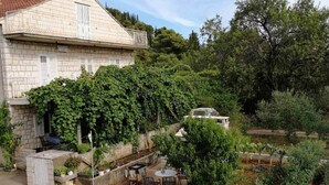Un edificio in pietra con un balcone, immerso nel verde e circondato da un giardino con diverse piante, un tavolino e sedie.