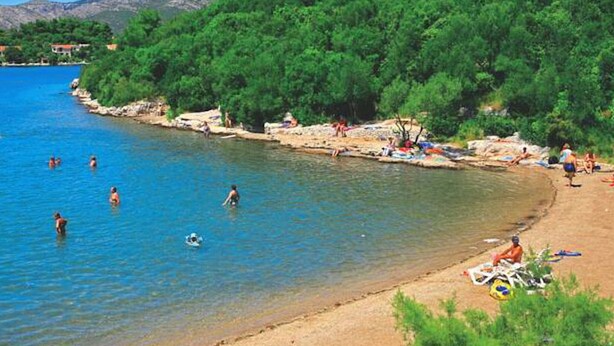 Una spiaggia con acque cristalline, persone che nuotano e si rilassano al sole, con montagne sullo sfondo.
